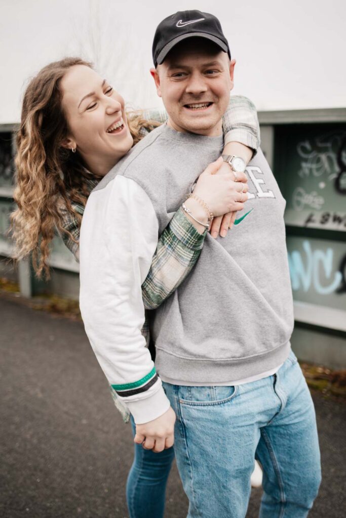 Paarshooting Phoenix West Dortmund Freundin umarmt Freund von hinten und lacht ihn an, Freund lacht in die Kamera