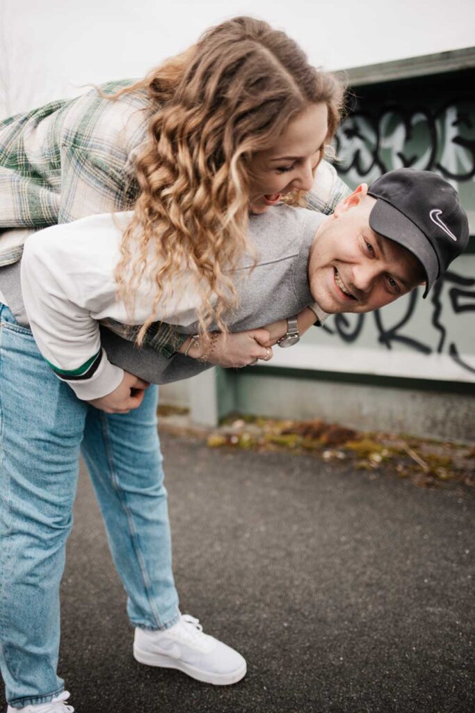 Paarshooting Phoenix West Dortmund Freund trägt Freundin Huckepack und lehnt sich stark nach vorne Freundin lächelt ihn an