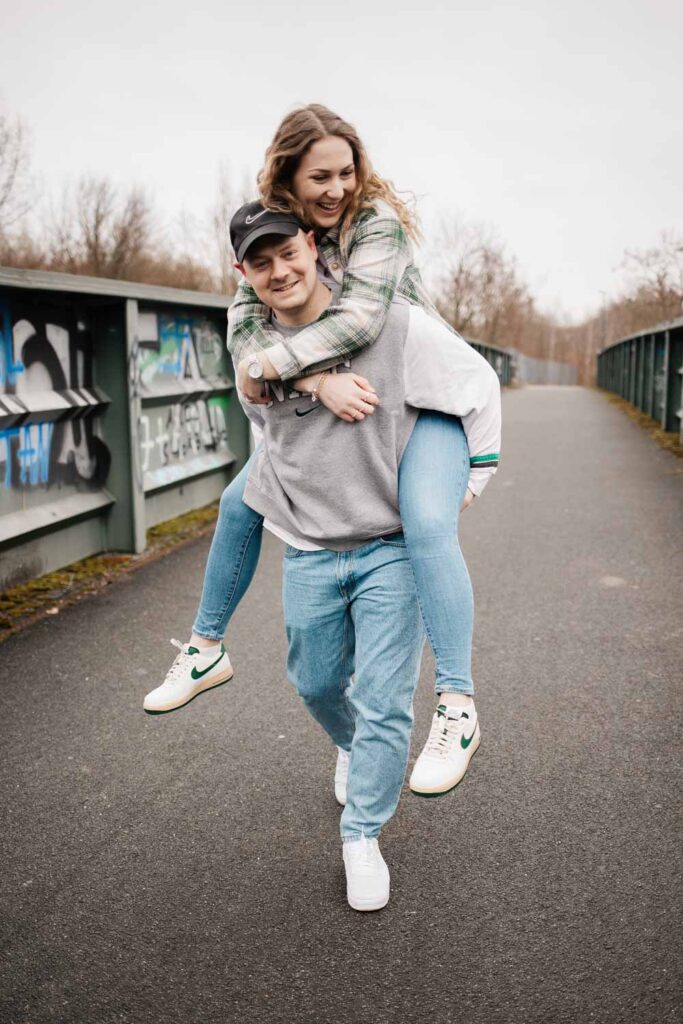 Paar läuft über die Brücke, Mann trägt Frau Huckepack, sie lachen dabei