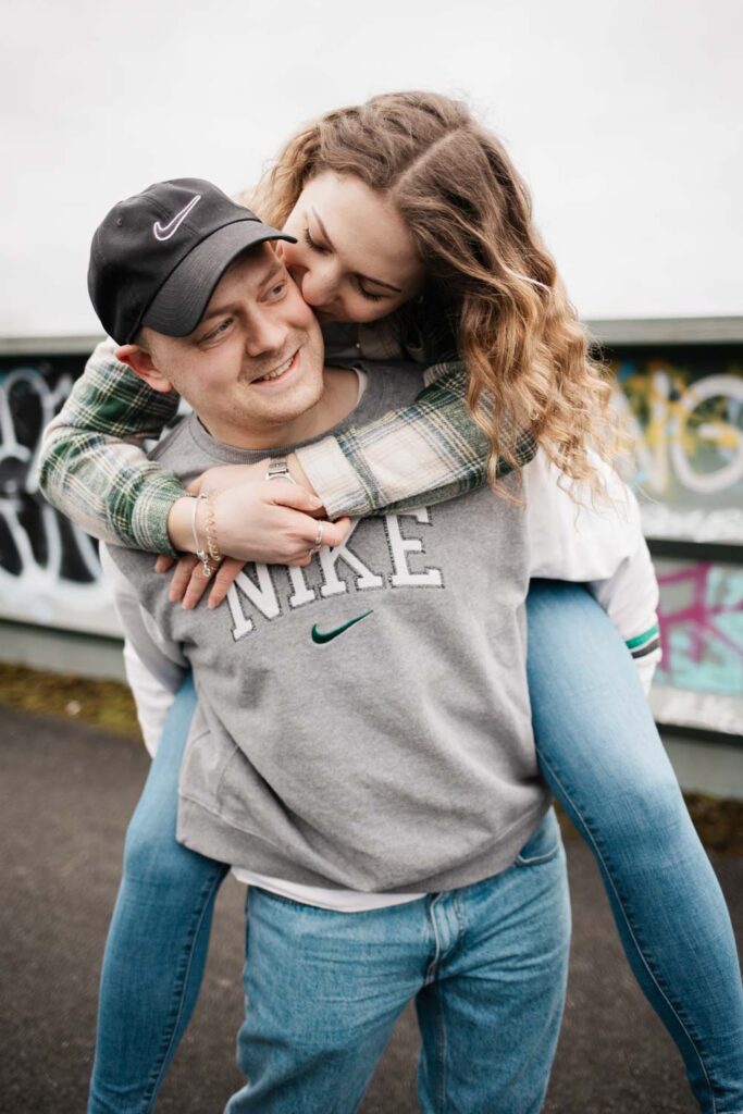 Paar läuft über die Brücke, Mann trägt Frau Huckepack, sie lachen dabei Freundin küsst Freund auf die Wange