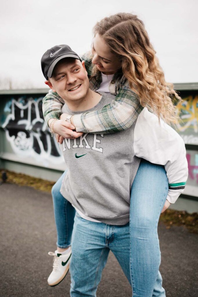 Paar läuft über die Brücke, Mann trägt Frau Huckepack, sie lachen dabei und Freundin schaut Freund an